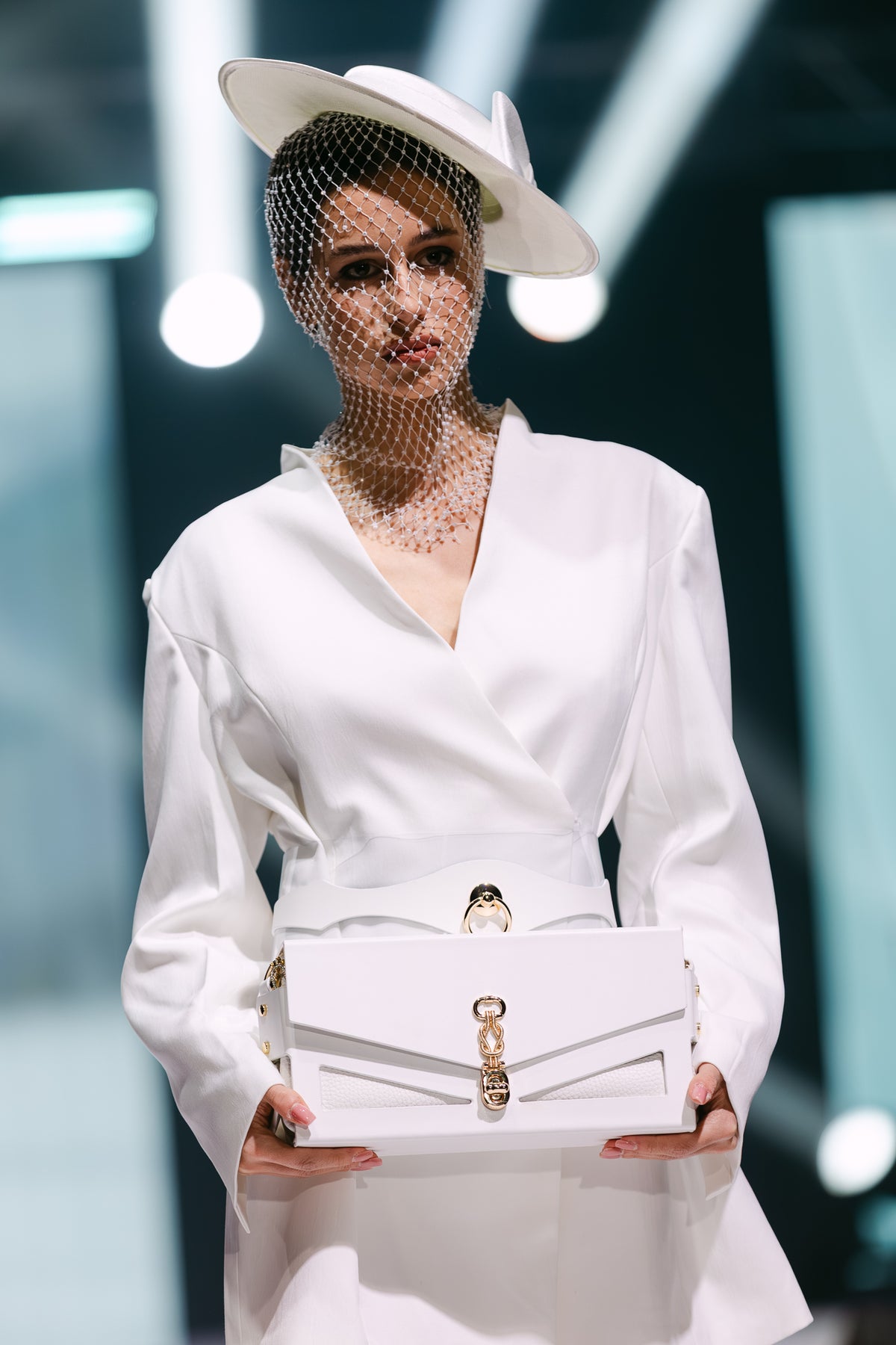 Model in white holds J’ÔTE white leather handbag with gold knot clasp on runway