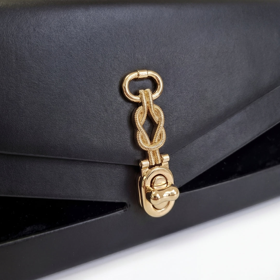 Nahaufnahme des goldenen Knoten-Signaturverschlusses der J’ÔTE Handtasche auf glattem schwarzem Leder